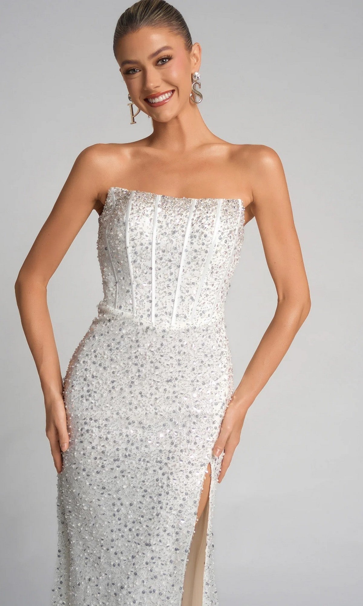 A woman poses and smiles in the Portia and Scarlett XO054 long formal dress, featuring a strapless corset bodice and sequined white skirt with a high slit, set against a plain background.